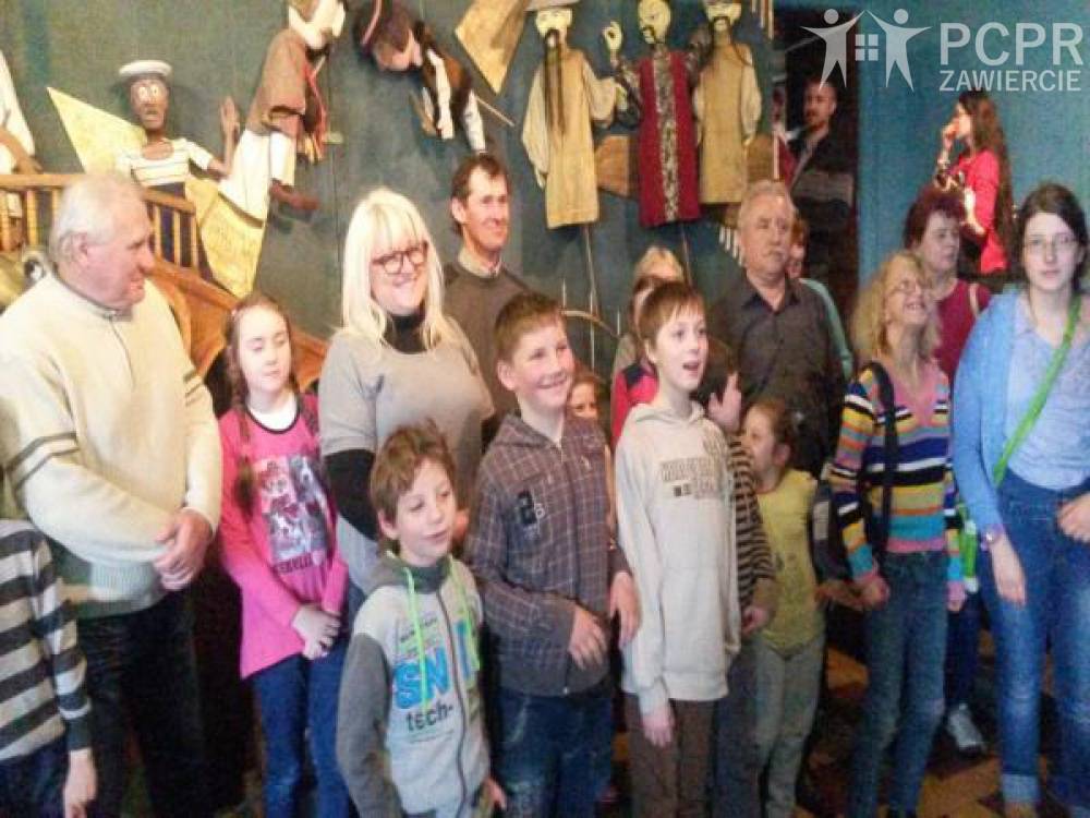 Zdjęcie: Grupa uczestników i uczestniczek projektu wraz z dziećmi stojąca we wnętrzu Teatru, a w tle kukiełki z przedstawień